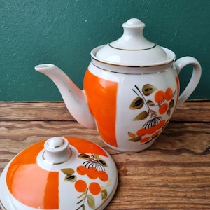 May include: A white teapot with an orange and white floral design. The teapot has a lid and a spout. The teapot is sitting on a wooden surface.