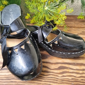 May include: Black patent leather Mary Jane style shoes with cut-out triangle details and black ribbon ties. The shoes have a glossy finish and a wooden sole. The brand name "a. nord" is visible on the insole.
