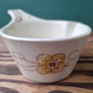 Op de afbeelding: Een witte keramische roomkan met een bruin en geel bloemenontwerp. De roomkan heeft een uniek handvat dat om de zijkant buigt.