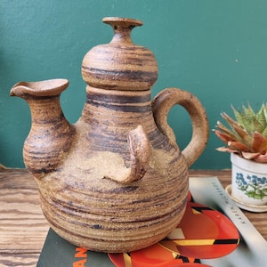 May include: A brown and black ceramic teapot with a lid and a handle. The teapot is sitting on a book titled "Art Deco and Modernist Ceramics".