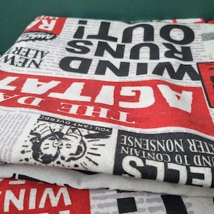 May include: Folded fabric with a newspaper print design. The print features bold red and black text, including phrases like "WIND RUNS OUT!" and "AGITATE." The fabric is white with a newsprint pattern.