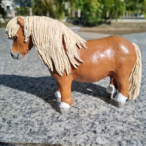 May include: A brown and white ceramic figurine of a horse with a long, flowing mane. The horse is standing on a gray surface.