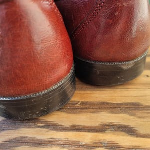 May include: A pair of burgundy leather loafers with black soles. The loafers are sitting on a wooden surface.