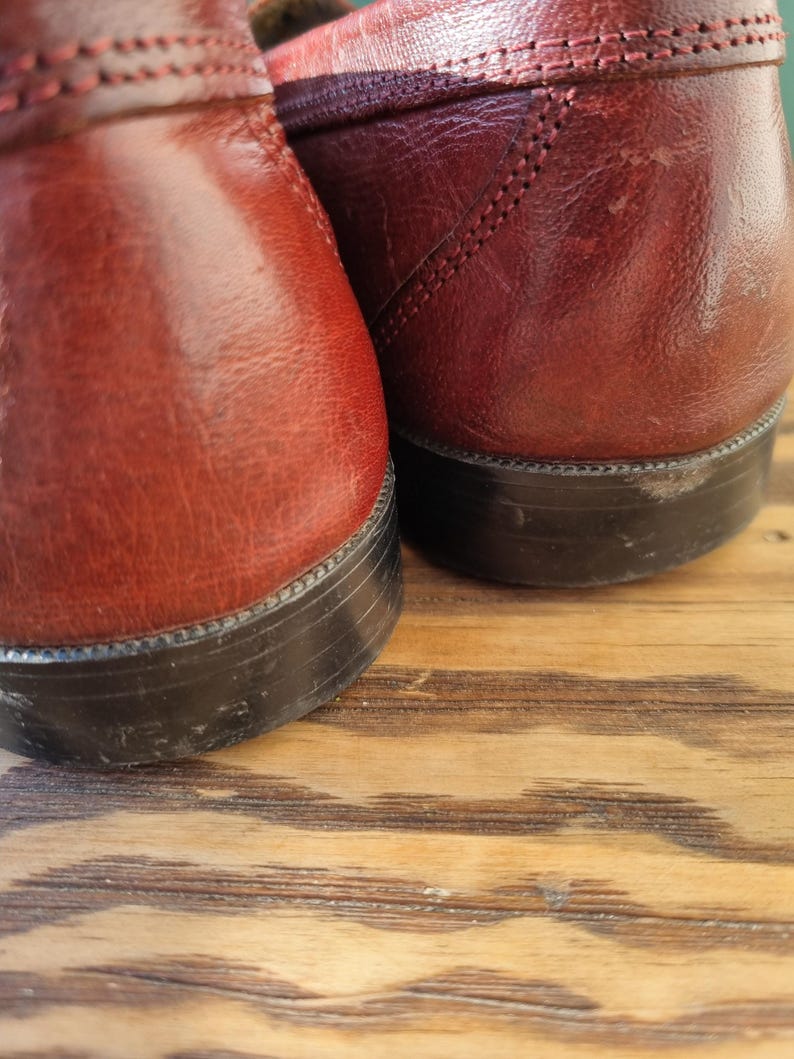 May include: A pair of burgundy leather loafers with black soles. The loafers are sitting on a wooden surface.