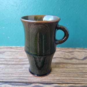 May include: A dark green ceramic mug with a handle. The mug has a flared top and a slightly textured surface. The mug is approximately 4 inches tall and sits on a wooden surface against a teal background.