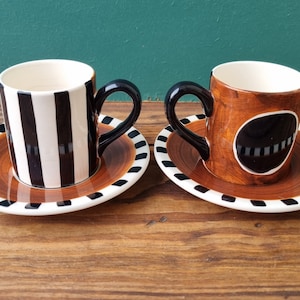 May include: Two ceramic coffee cups with matching saucers. One cup has black and white stripes, the other has a brown and black swirl design with a black circle in the center.