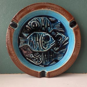 May include: A ceramic ashtray with a brown rim and a turquoise interior. The centre features a raised design of three stylized fish in shades of blue. The ashtray has three small notches for resting cigarettes.