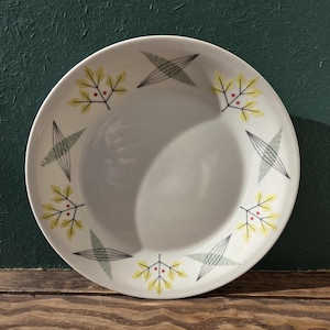 May include: White ceramic plate with a mid-century modern design. The plate features a repeating pattern of yellow leaves with red berries and gray geometric shapes around the rim. The plate is set against a dark green background.