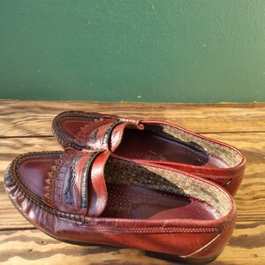 May include: A pair of burgundy leather loafers with a decorative stitch pattern on the toe. The loafers have a black leather strap across the top and a brown leather lining.