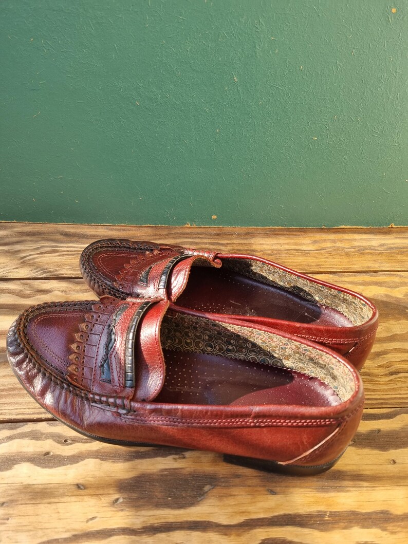May include: A pair of burgundy leather loafers with a decorative stitch pattern on the toe. The loafers have a black leather strap across the top and a brown leather lining.