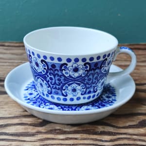 May include: A white teacup and saucer set with a blue floral pattern. The cup has a handle and sits on a matching saucer. The blue design includes flowers and swirls. The set is on a wooden surface.
