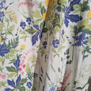 May include: A white fabric with a floral pattern featuring blue, yellow, pink, and green flowers. The fabric has a linen-like texture.