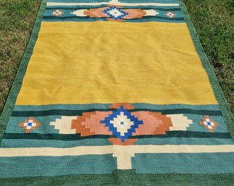 May include: A rectangular woven rug with a yellow center and geometric patterns in teal, orange, and white. The rug has a green border and white fringe along the short edges. The design features diamond and cross shapes.