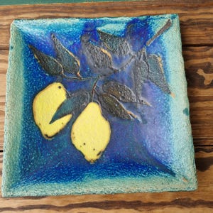 Puede incluir: Un plato cuadrado de cerámica con un esmalte azul y verde. El plato presenta un diseño pintado de una rama de limonero con dos limones amarillos.