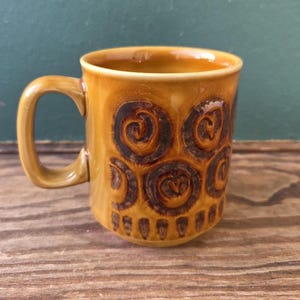 Può includere: Una tazza in ceramica con una glassa marrone e un motivo a spirale marrone. La tazza ha un manico curvo e una forma cilindrica. Il disegno si ripete attorno all'esterno della tazza. La tazza è appoggiata su una superficie di legno.