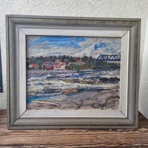 May include: An oil painting in a silver-toned frame depicts a landscape with a river, bridge, and buildings under a cloudy blue sky. The artwork uses textured brushstrokes to create depth and movement in the water and sky.