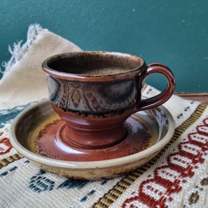 Peut inclure: Un ensemble tasse et soucoupe à café en céramique. La tasse est marron foncé avec un intérieur marron clair et une anse arrondie. La soucoupe est de couleur assortie avec un centre légèrement surélevé. L'ensemble est posé sur un textile à motifs.