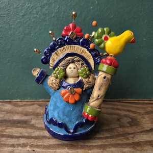 May include: A ceramic figurine featuring a central figure with light brown hair, wearing a blue dress with an orange flower accent. The figure has a dark blue headdress with a red flower and a yellow bird perched on a textured tan staff.