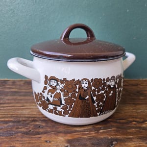 Puede incluir: Una olla de esmalte blanco con tapa marrón oscuro y dos asas laterales. El exterior presenta un estampado gráfico marrón de tres figuras con ropa de estilo medieval rodeadas de grandes flores y hojas estilizadas sobre un fondo blanco.