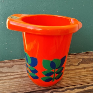 May include: Bright orange plastic planter with a unique handle design. The planter features a repeating pattern of blue and green stylized leaves. The planter is sitting on a wooden surface.