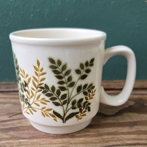 May include: White ceramic mug with a curved handle, featuring a botanical design of brown and gold leaves. The mug has a stepped base and a simple, classic shape, suitable for hot beverages.