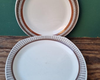 May include: Two white ceramic plates with brown and orange geometric patterns around the rim. The plate on top has a round shape, while the plate on the bottom has a slightly oval shape.