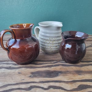 May include: Three ceramic pitchers in varying sizes and colors sit on a wooden surface. The largest is a glossy brown with a handle. The middle pitcher is white with a speckled pattern. The smallest is a dark brown with a textured design.