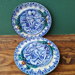 Könnte beinhalten: Zwei blaue und weiße Keramikteller mit einem erhabenen Design von Figuren in einem Kreis. Die Figuren sind in Blau und Weiß und sind von grünen Blättern und roten Blumen umgeben.