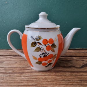 Puede incluir: Una tetera de porcelana blanca con ribete dorado y diseño floral naranja. La tetera tiene tapa y asa.
