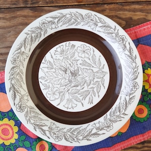 Puede incluir: Un plato redondo blanco con centro y borde marrón. El plato presenta un diseño floral en marrón y blanco. El patrón floral incluye hojas y flores. El plato está sobre un paño estampado colorido.