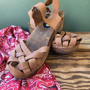 May include: A pair of brown leather sandals with a woven design on the front and a wooden sole. The sandals have a strap that goes around the ankle. A red and white patterned fabric is in the background.