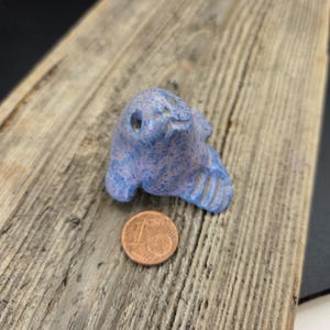 May include: A small, blue ceramic seal figurine with a speckled pattern. The seal has a smiling face and flipper details. A one-cent coin is next to the seal for size comparison. The figurine is on a weathered wooden surface.