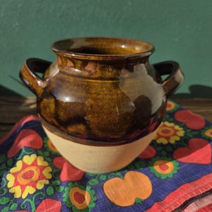 May include: A handcrafted ceramic pot with two handles, featuring a glossy brown glaze over a cream-colored base. The pot sits on a colorful patterned cloth with floral and heart motifs, against a green backdrop.