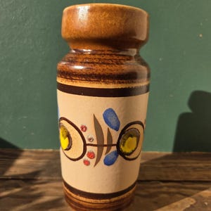 May include: A brown ceramic vase with a cylindrical shape and a slightly flared top. The vase features a tan band with a decorative pattern of yellow, blue, and red accents. The vase is approximately 6 inches tall.
