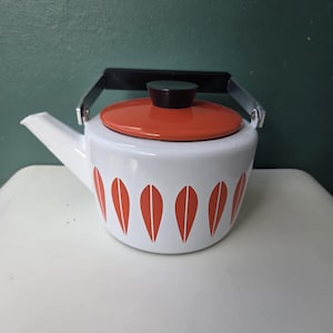May include: White enamel teapot with an orange lid and a black handle. The teapot has a pattern of orange leaf shapes with white accents. The spout is white, and the handle is black. The teapot is on a white surface.