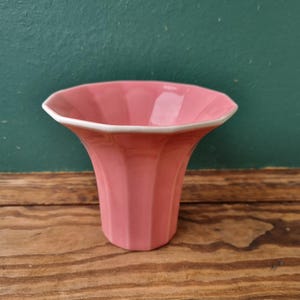 Puede incluir: Un jarrón de cerámica rosa con un diseño acanalado. El jarrón tiene un borde blanco y está sobre una superficie de madera.