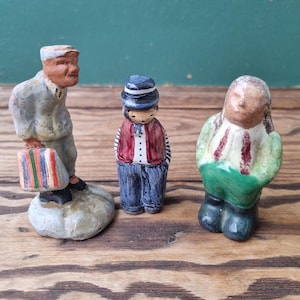 Peut inclure: Trois petites figurines en céramique peintes à la main. L'une tient une valise à rayures colorées. Une autre porte une veste rouge et un pantalon bleu. La troisième porte une tenue verte et blanche.