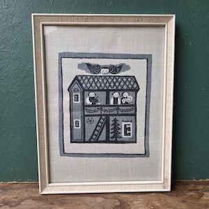 May include: Framed vintage folk art textile featuring a two-story house with figures in the windows and on the balcony. The artwork is primarily in shades of gray and white, with a decorative border and a light-colored wooden frame.