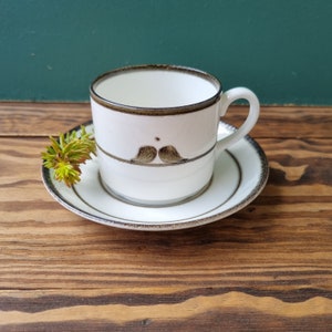 Puede incluir: Una taza de café de cerámica blanca con un borde marrón y un diseño de pájaro marrón. La taza está sentada sobre un platillo a juego.