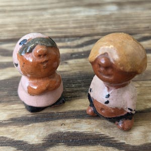 May include: Two small, hand-painted ceramic figurines. One has a light brown head and a pink body, the other has a tan head and a pink body. Both have dark brown facial features and black accents. They are standing on a wooden surface.