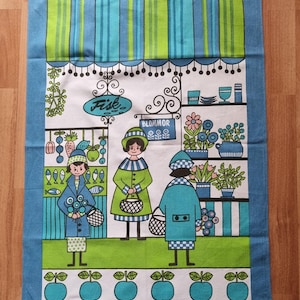 Puede incluir: Una toalla de cocina vintage con un diseño azul, verde y blanco. La toalla presenta una ilustración de dibujos animados de un mercado con tres personas comprando pescado, flores y fruta. El texto "Fisk" y "Blommor" es visible en la toalla.