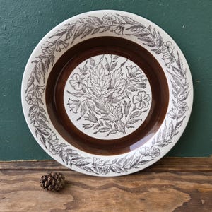 Puede incluir: Un plato redondo de cerámica vintage con un diseño floral marrón y blanco. El plato presenta un anillo central marrón y un borde de hojas y flores. Una pequeña piña se encuentra frente al plato sobre una superficie de madera.