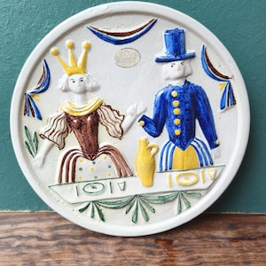 May include: A white ceramic decorative plate with a relief design of a royal couple in period attire. A yellow jug sits between them. The plate has a scalloped edge and the text "Mills" is visible. The plate is likely from the Mills brand.