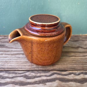 Könnte beinhalten: Eine kleine, runde Keramik-Teekanne mit einer glänzenden, gesprenkelten bernsteinbraunen Glasur. Die Teekanne verfügt über einen kurzen, geschwungenen Ausguss, einen stabilen Seitengriff und einen passenden runden Deckel mit dunklerem Rand.