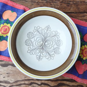 Puede incluir: Un cuenco redondo de cerámica blanca con borde marrón y amarillo. El cuenco presenta un diseño floral gris y ondulado en el centro. El cuenco está sobre una superficie de madera con un paño estampado colorido.