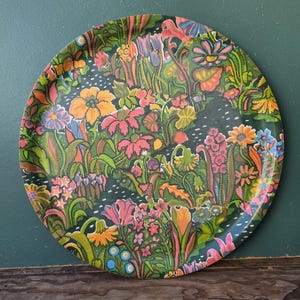 May include: Circular serving tray with a colourful floral pattern. The tray has a dark green base, adorned with stylised flowers in orange, pink, yellow, and blue. The flowers are densely packed, creating a lively and decorative surface, perfect for serving food or drinks.