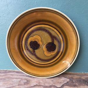 May include: A round ceramic plate featuring a glossy mustard yellow and brown glaze. The center displays a circular motif containing two stylized flower shapes in dark brown and yellow, surrounded by concentric brown rings and a cream-colored rim.