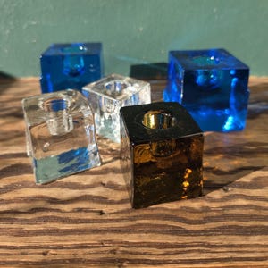 May include: A collection of square glass candle holders in various colors, including clear, blue, and amber. The candle holders are arranged on a wooden surface, showcasing their geometric design and transparency.