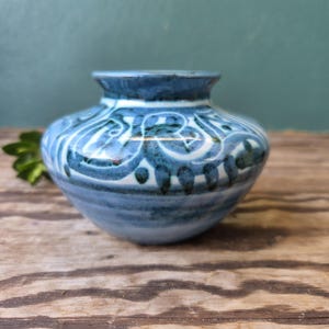 Pode incluir: Um pequeno vaso de cerâmica atarracado com corpo arredondado e pescoço curto e alargado. A superfície é decorada com padrões de redemoinhos azuis e brancos pintados à mão, com detalhes em azul escuro criando uma aparência texturizada.
