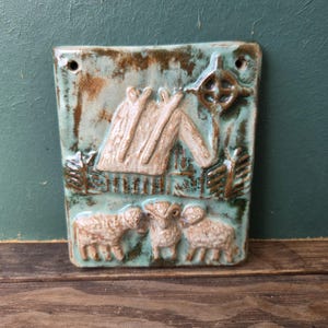 Op de afbeelding: Rechthoekige keramische wanddecoratie met een turquoise glazuur. Het reliëfontwerp toont een tent, een zonnesymbool, bomen en drie schapen. Het stuk heeft twee gaten aan de bovenkant om op te hangen. De algehele stijl is rustiek en artistiek.